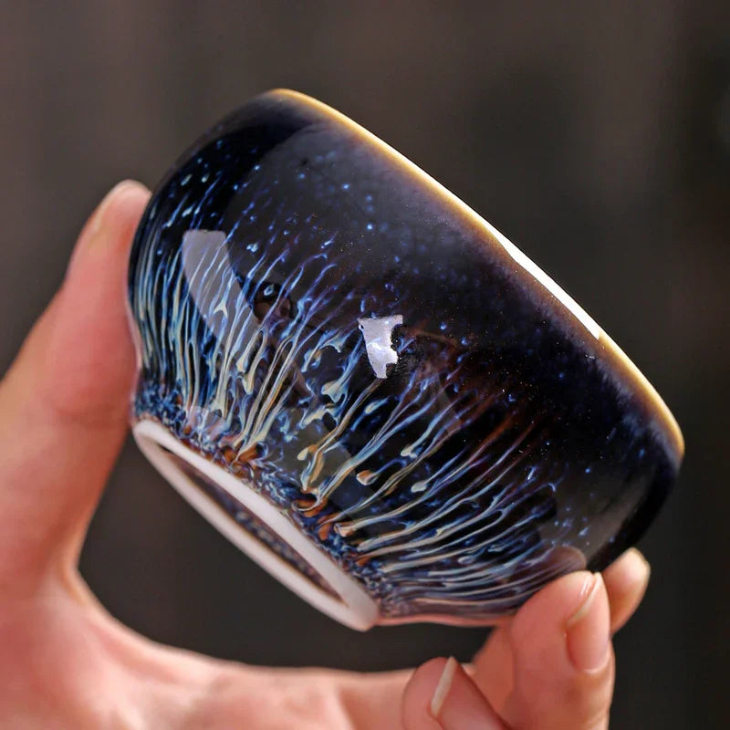 Hand holding a dark blue ceramic tea bowl with a textured, glossy glaze and drip patterns