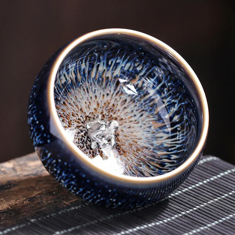 Close-up of a ceramic bowl with intricate blue and brown glaze and a silver frog figurine inside