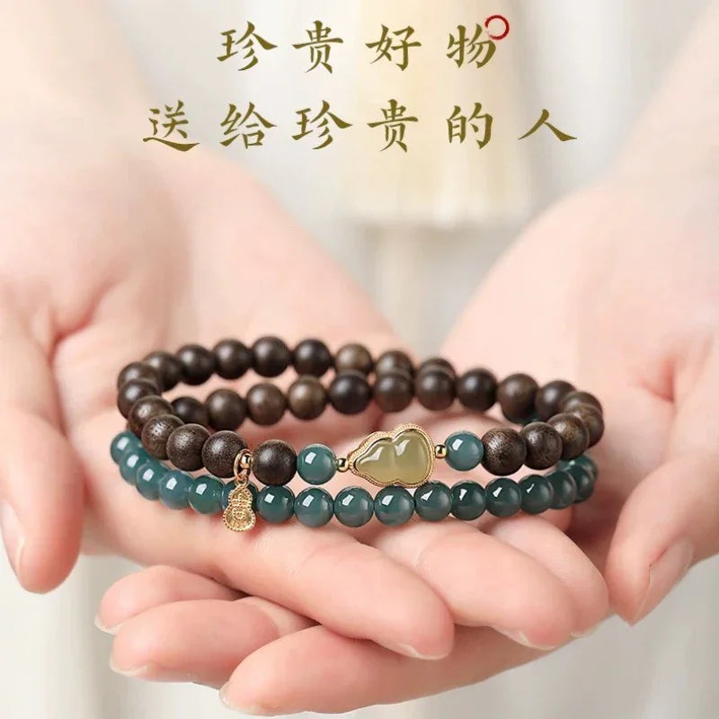 Hand holding two beaded bracelets, one with dark wood beads and gold charm, the other with green gemstone beads