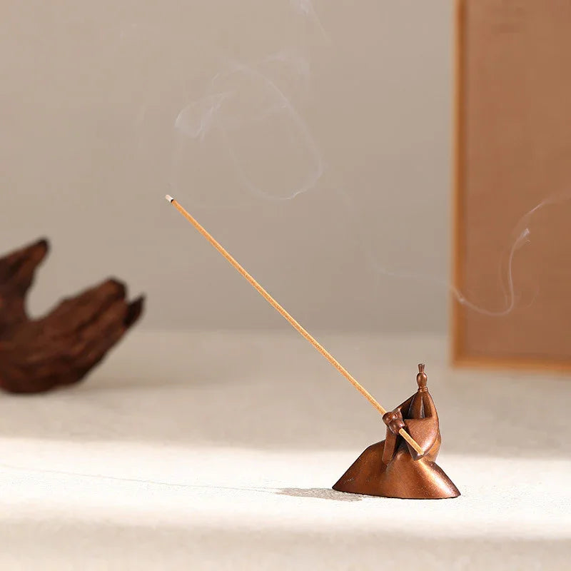 Bronze incense holder shaped like a seated figure holding a long incense stick with smoke curling up