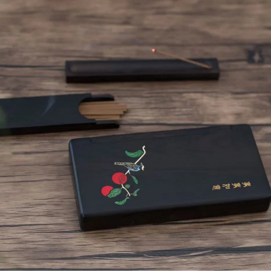 Black incense holder box with bird and red fruit design on wooden surface, open with incense sticks and lit stick