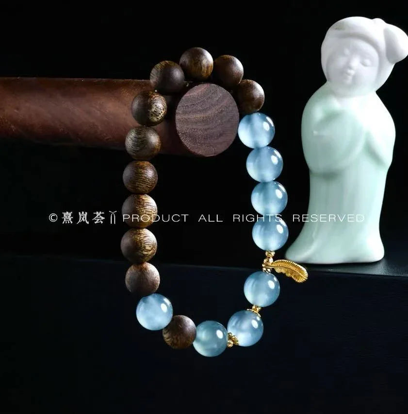 Wood and blue glass bead bracelet with gold feather charm displayed with white Buddha statue