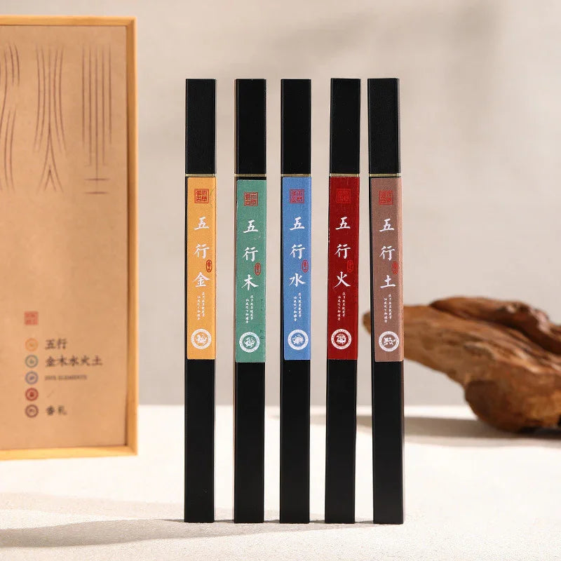 Set of five black incense sticks with colored labels representing five elements, next to wooden box and driftwood