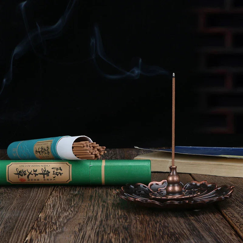 Burning incense stick in metal lotus holder with green incense box on wooden table and open book