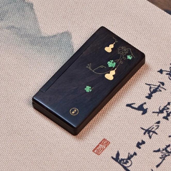 Black wooden rectangular ink stone with gold and green gourd vine inlay on beige fabric with Chinese calligraphy