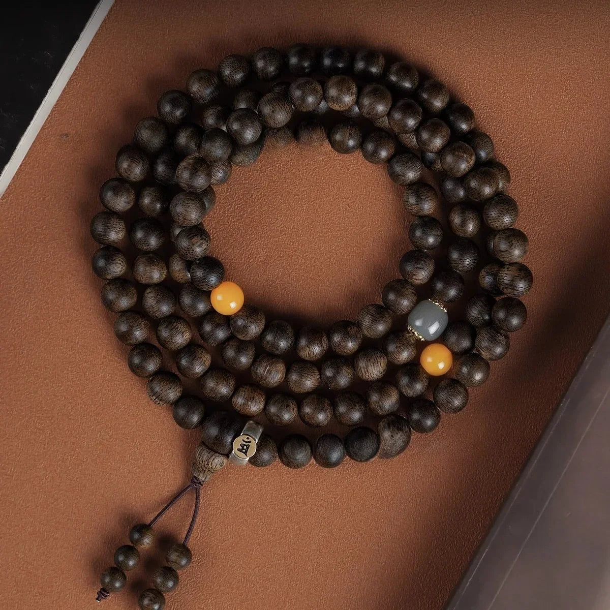 Dark brown wooden prayer beads with two orange and one gray bead, lying coiled on a brown surface
