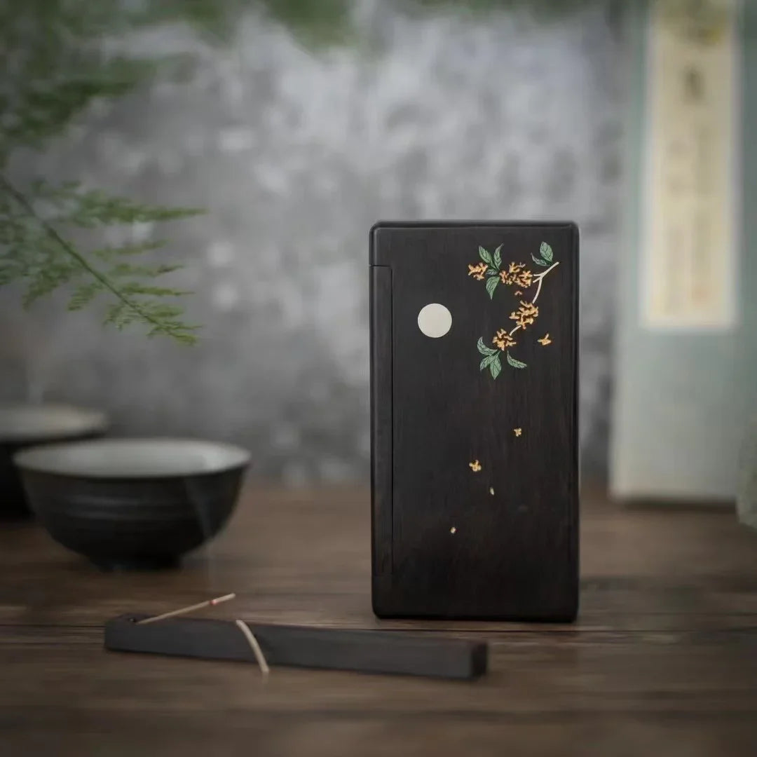 Black wooden incense box with floral design and moon illustration on wooden table with ceramic bowls