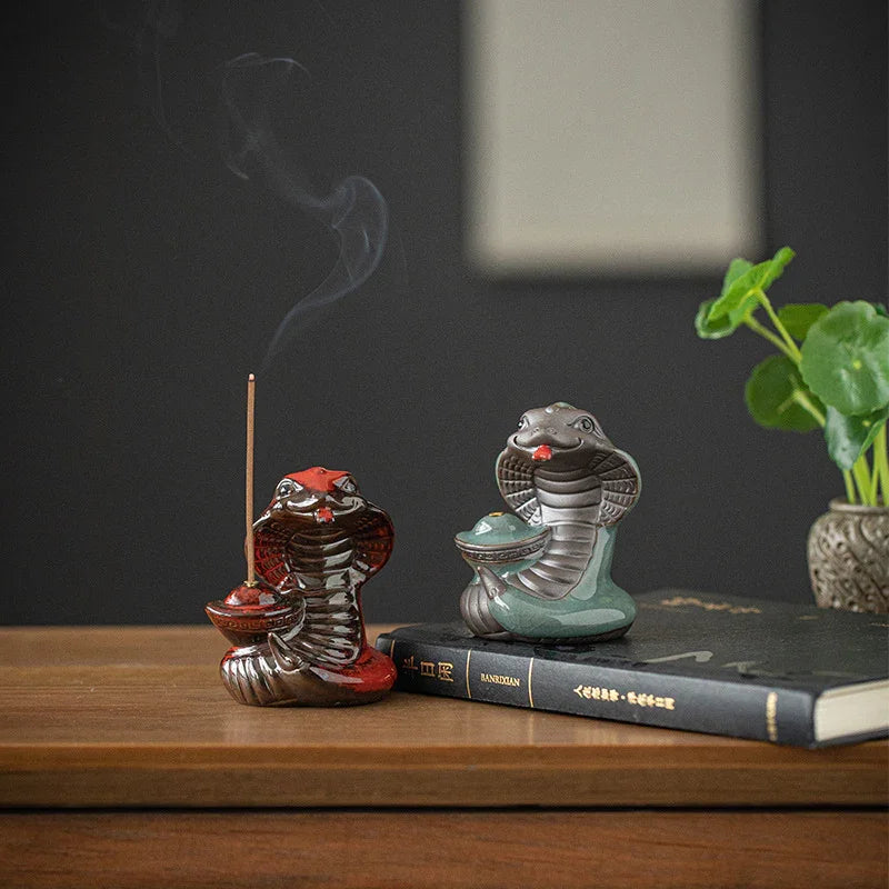 Red and green ceramic cobra incense holders on wooden table with smoke, book and green plant